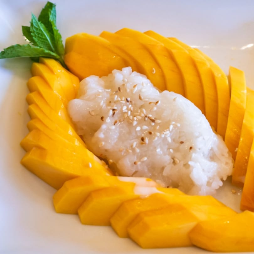 Thai mango sticky rice dessert with sesame seeds and a mint garnish on a white plate.