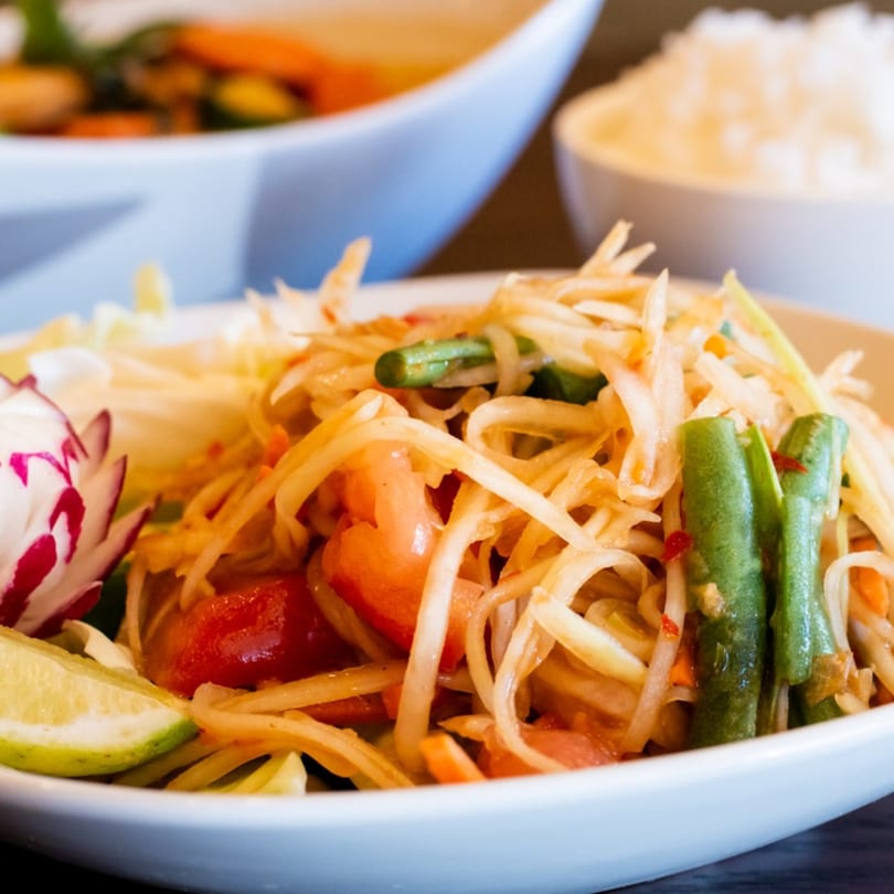 Close-up of Thai papaya salad with green beans, tomatoes, and lime in a white bowl.